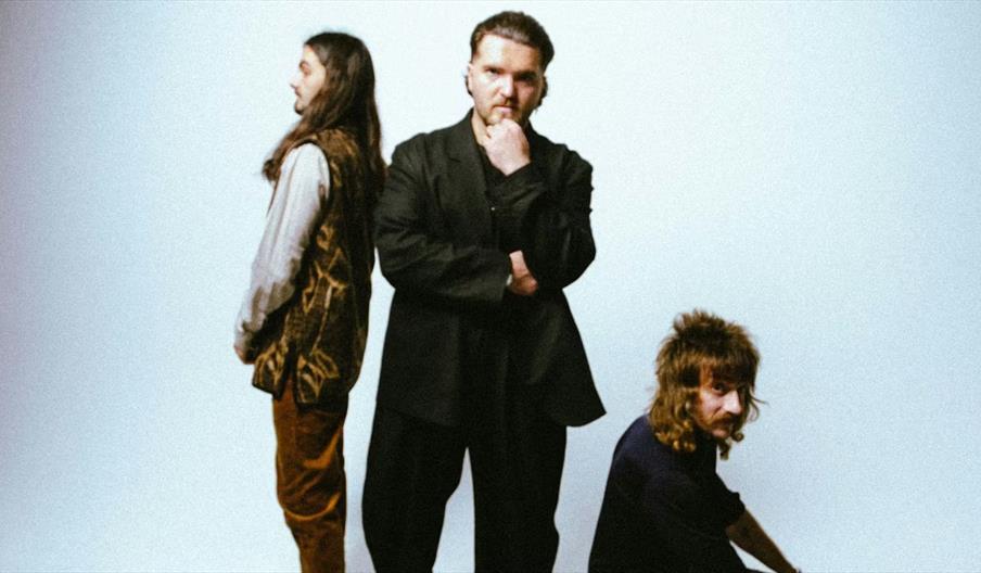 Three men pose against a light background; one stands in the center with his hand on his chin, another stands to the left facing sideways, and the thi