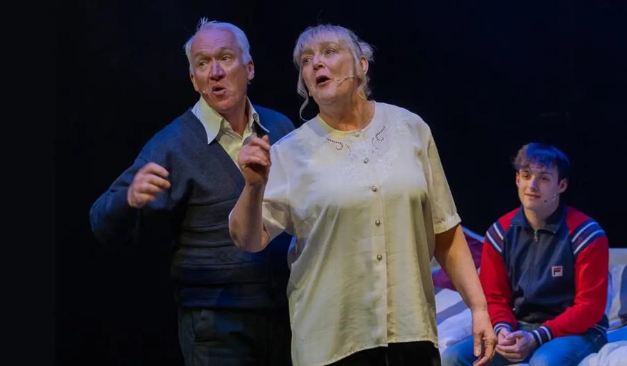 Two older adults, a man and a woman, are standing and singing or speaking on stage with microphones, while a younger man in a red and blue jacket sits