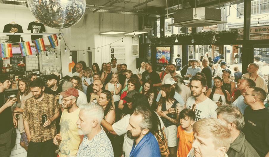 A large and diverse crowd gathers inside Humber Street Gallery, standing attentively under a disco ball with rainbow flags hanging above the bar, whil