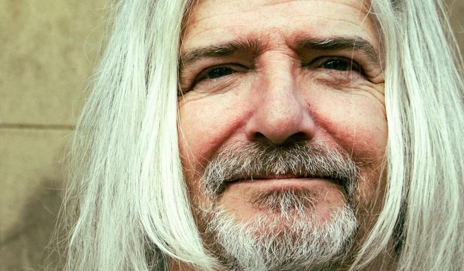 Close-up of an older man with long white hair and a white beard, wearing a brown shirt and smiling slightly, set against a plain tan background.