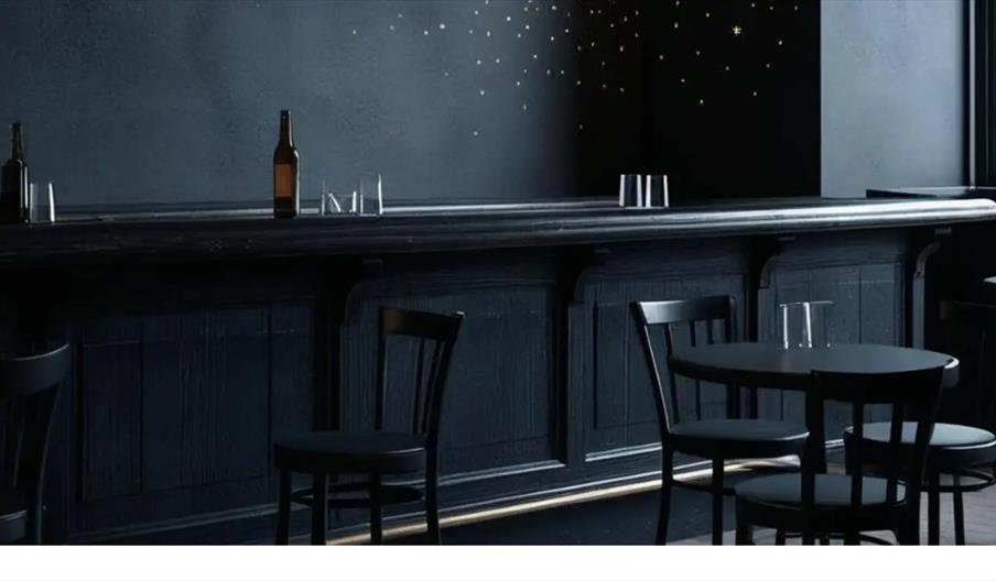 A dimly lit bar with black furniture, including a bar counter and several chairs and tables, features a few empty glasses and beer bottles on the coun