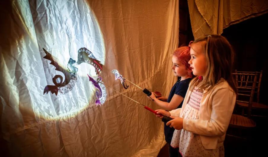 Two young girls stand side by side, holding sticks with dragon-shaped shadow puppets, projecting their shadows onto a white sheet illuminated by a bri