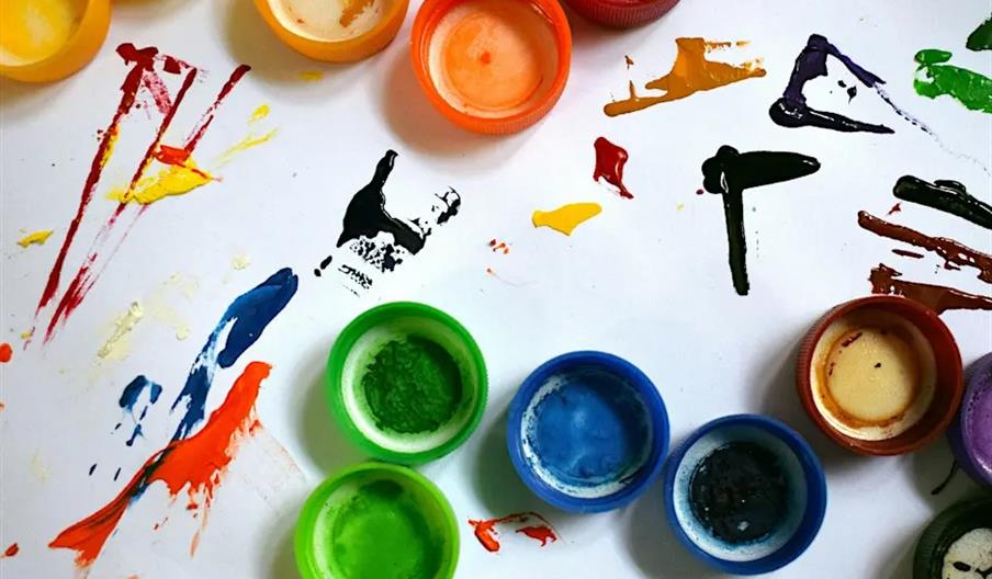 Plastic paint containers in various colors are arranged on a white surface, which is covered with colorful paint splatters and brush strokes in red, y