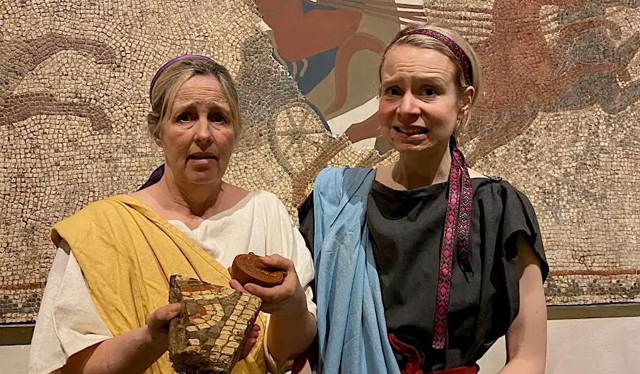 Two women dressed in ancient Roman-style clothing stand in front of a mosaic wall depicting a chariot scene; one woman holds a piece of mosaic and a r