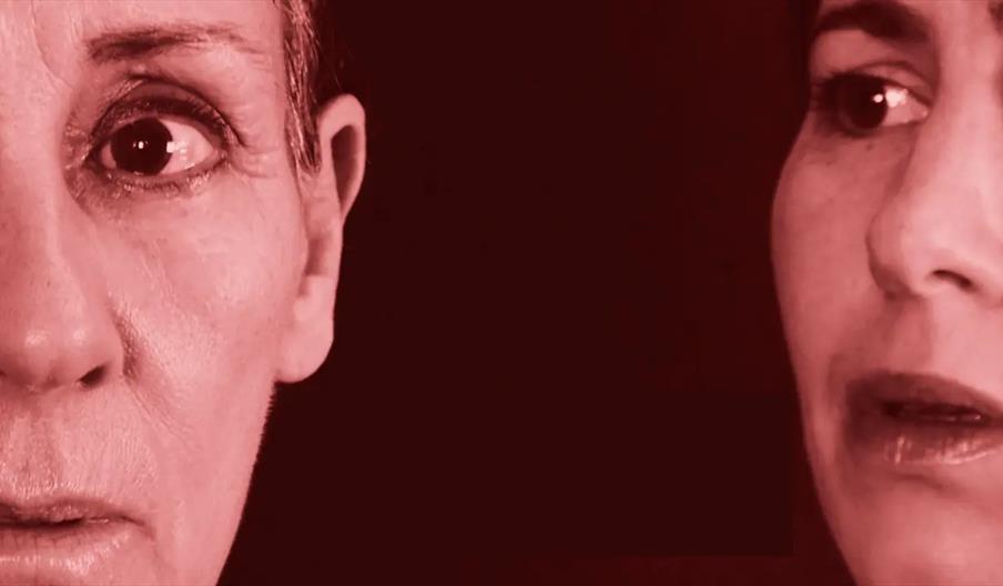 Close-up image of two human faces in red-tinted lighting, with one person on the left facing forward and the other on the right, both cropped so only