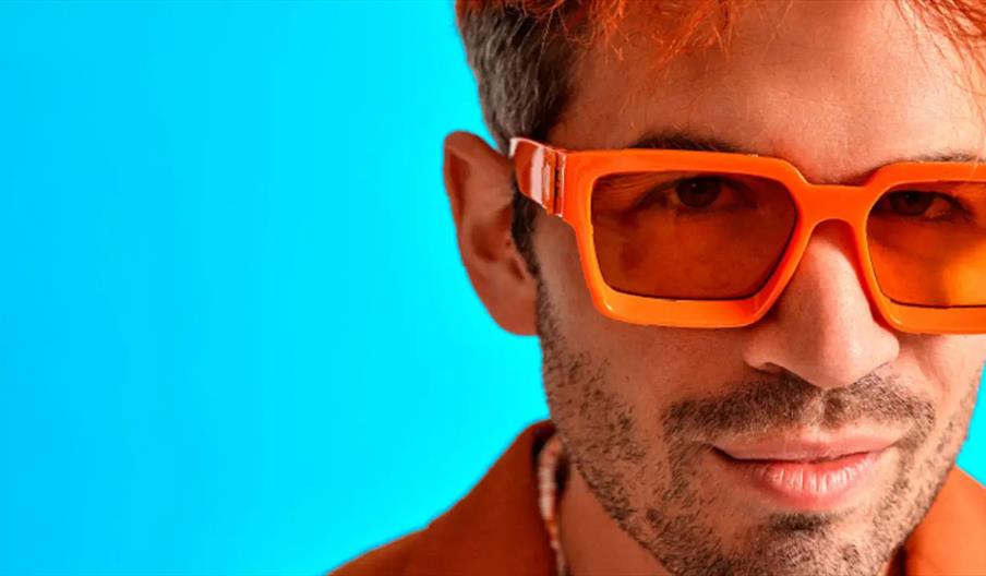 Close-up of a person wearing large, bright orange sunglasses and an orange shirt, set against a vibrant blue background. Only the right side of the pe