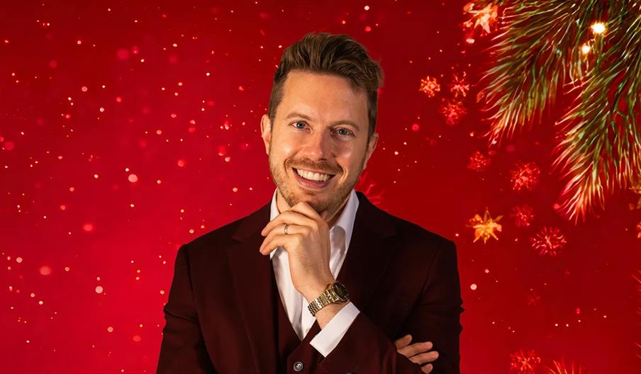A smiling man in a maroon suit poses with his hand on his chin in front of a festive red background with sparkling lights, snowflake shapes, and pine