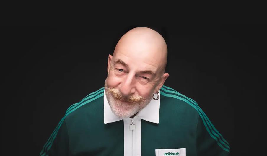 Smiling bald man with a bushy mustache and beard, wearing a green Adidas track jacket, poses against a plain black background.
