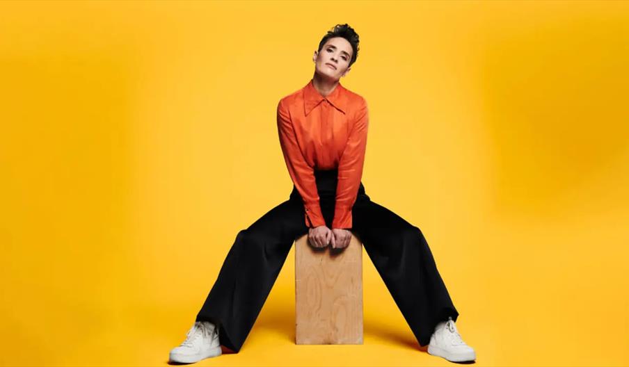 A person wearing a bright orange shirt, black wide-leg pants, and white sneakers sits on a wooden block against a solid yellow background, with their