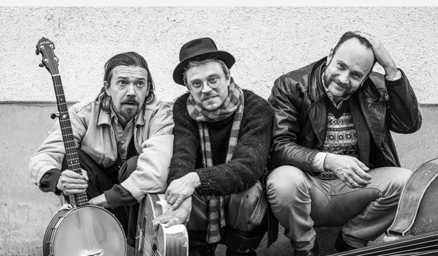 Black and white photo of three men sitting on the ground against a wall, each with a musical instrument (a banjo, a guitar, and a double bass). Large