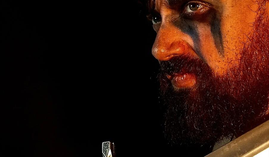 A close-up of a man with a thick beard and dramatic black face paint under his eyes, holding a large sword upright against his face, set against a dar