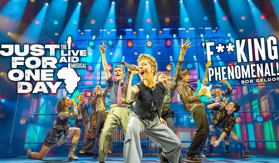 A group of energetic performers sing and dance on a brightly lit stage with colourful lights, advertising "Just For One Day: The Live Aid Musical." La