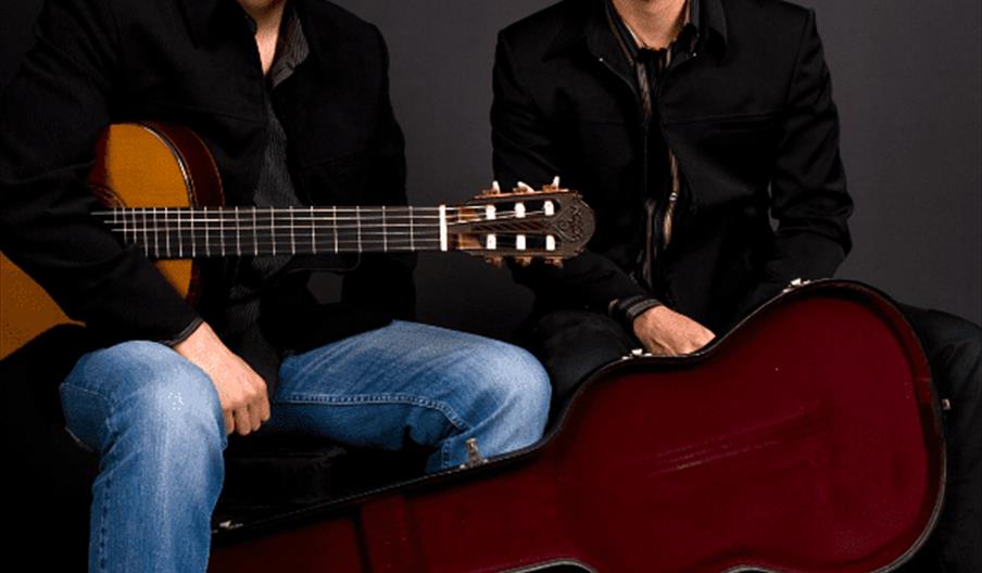 Two men in black jackets sit side by side against a dark background; one holds an acoustic guitar while the other sits next to a guitar in an open red