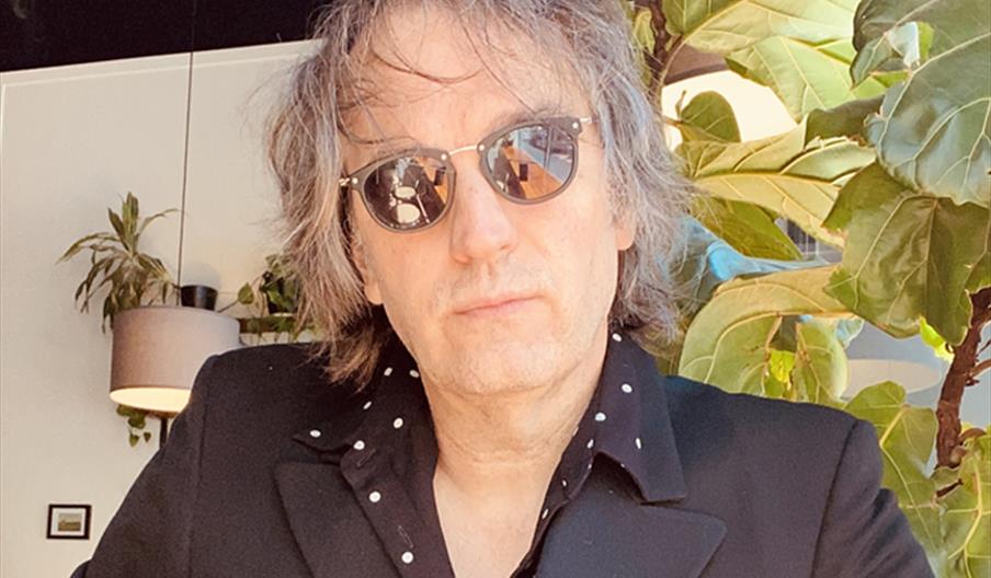 A person with tousled hair and sunglasses is wearing a black jacket over a black shirt with small white dots, seated indoors near a large leafy plant,