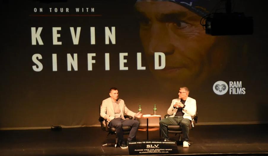 Two men are seated on stage in armchairs having a discussion, with a small table and water bottles between them; behind them, a large projection reads