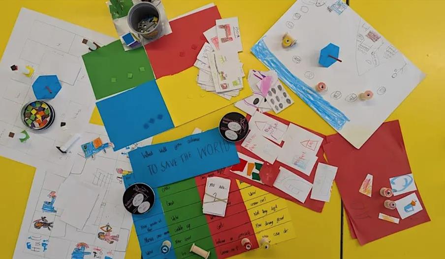 A variety of colourful papers, hand-drawn diagrams, cards, and small craft objects are spread out on a yellow table, suggesting a collaborative or cre