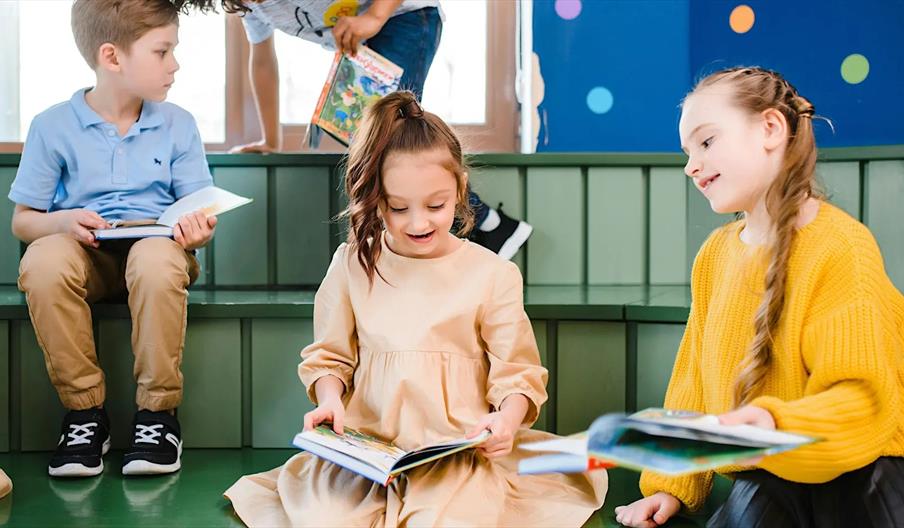Four young children are sitting on green steps, reading colorful books together in a brightly decorated room with blue and orange circles on the wall.