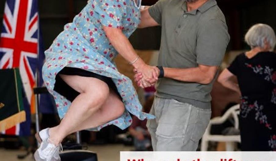 A woman in a blue polka-dot dress is airborne, smiling as she swings with a man in a grey shirt and beige pants, who supports her with one hand. They