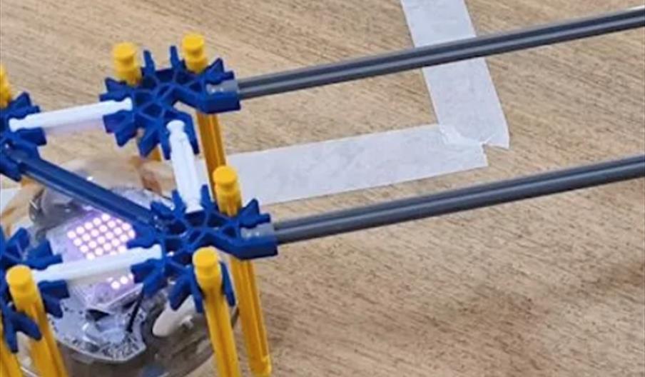 A small clear robot with colored lights and a frame built from blue, yellow, and white K'NEX pieces moves along a wooden surface, following white tape