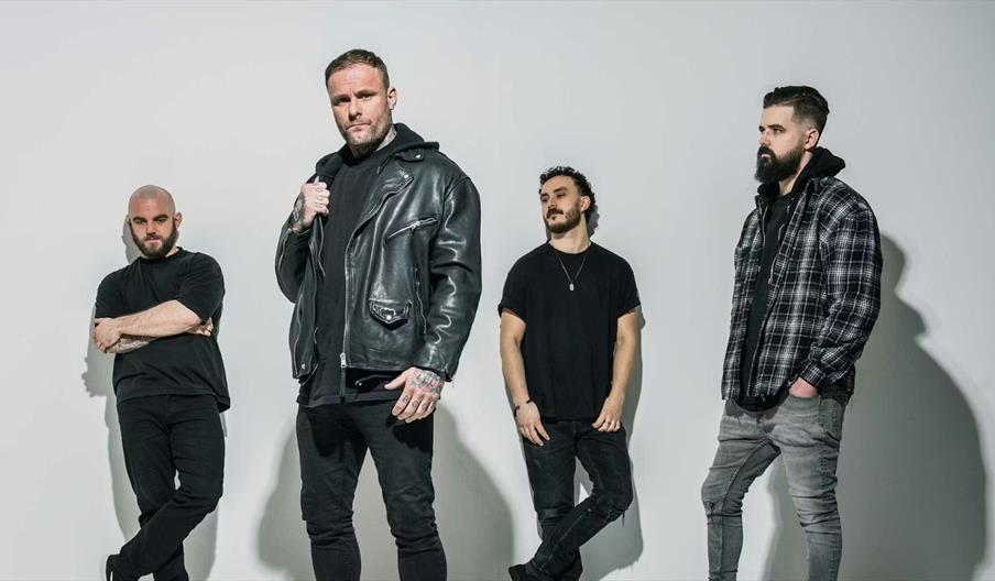 Four men from the Kris Barras Band stand against a plain light-colored background, dressed in casual dark clothing; the man second from the left wears
