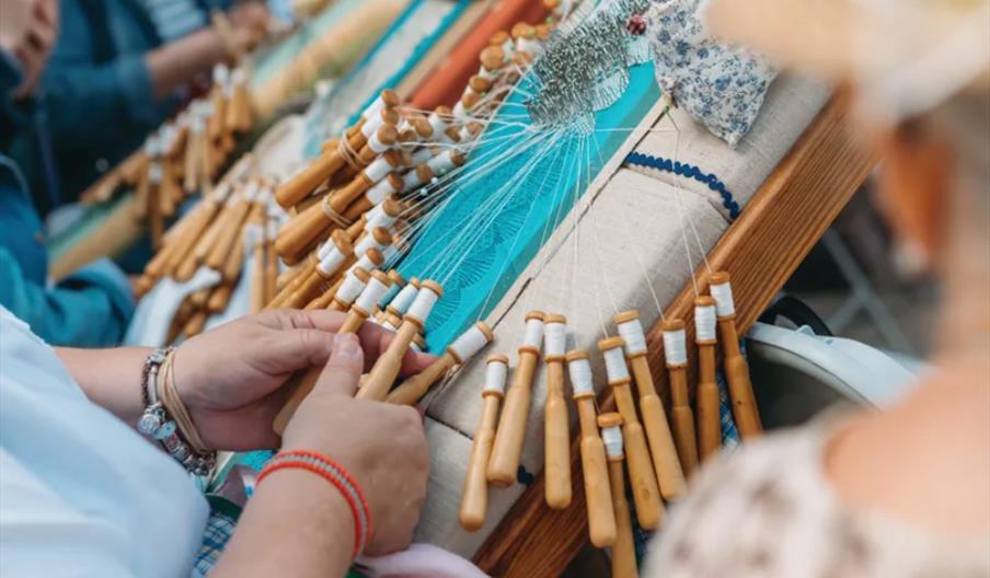 Close-up of hands making bobbin lace on a pillow with multiple wooden bobbins and white thread, at a craft demonstration, with other people working in