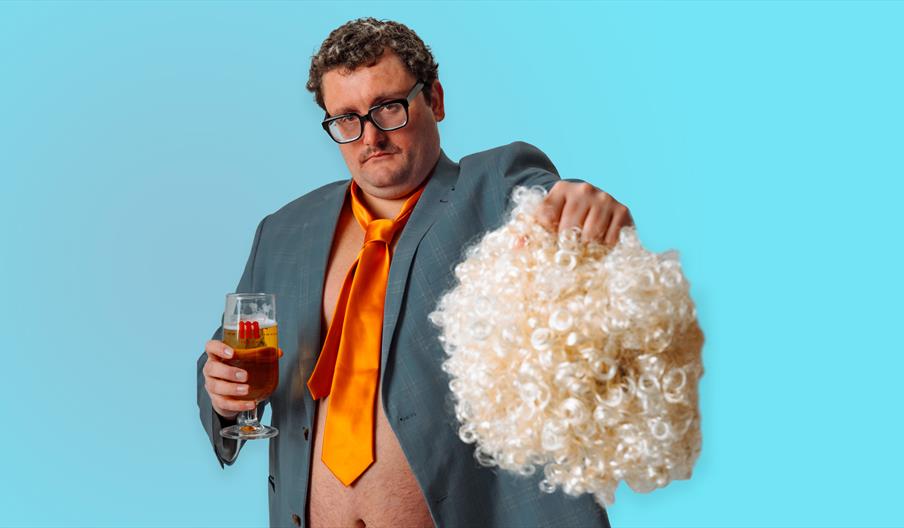 A man wearing a gray suit with an orange tie, unbuttoned to reveal his stomach, holds a glass of beer in one hand and a large, curly blond wig in the