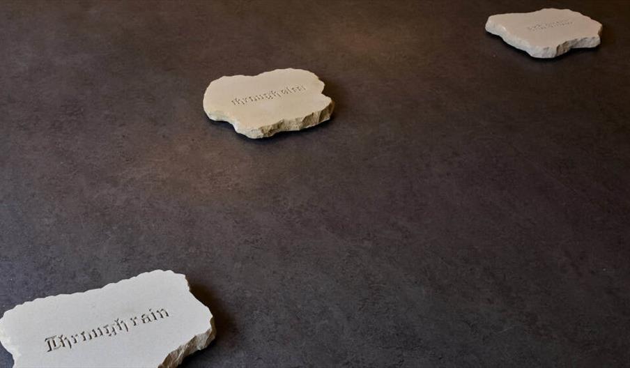 Three irregularly shaped stone slabs are placed on a dark floor, each engraved with a word in gothic-style lettering.