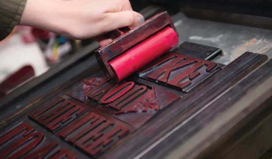 A person uses a roller with red ink to coat raised wooden letterpress type, preparing it for printing at the Ferens Art Gallery.