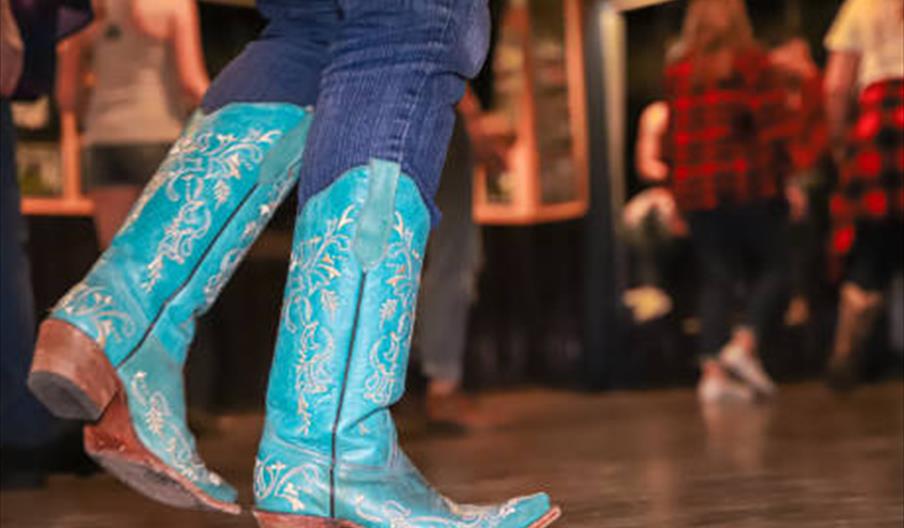 A close-up of a person wearing bright turquoise cowboy boots with white embroidery, dancing indoors on a smooth floor, with several other people blurr