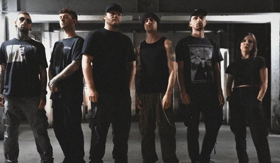 Six band members stand in a dimly lit, industrial setting, all dressed in dark, casual clothing and looking seriously at the camera. The group stands