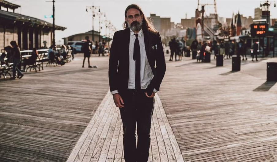 A man with long hair and a beard, dressed in a suit and tie, walks alone on a wooden boardwalk in the late afternoon, with groups of people and city b