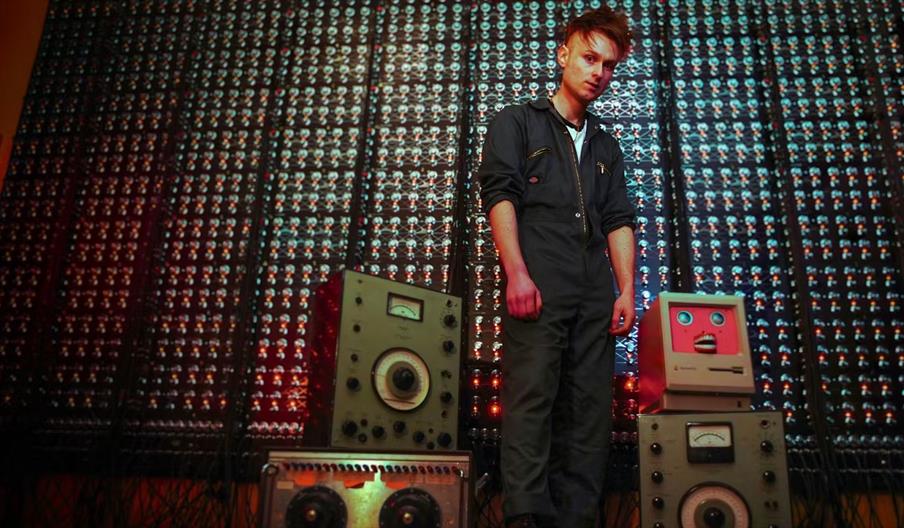 A person in a dark jumpsuit stands in front of a large wall of illuminated electronic components, with vintage computer and radio equipment, including