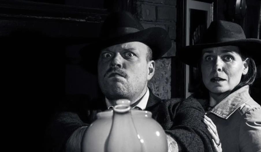 A black-and-white image shows a man and a woman wearing wide-brimmed hats looking intensely ahead; the man is in the foreground holding a large jug or