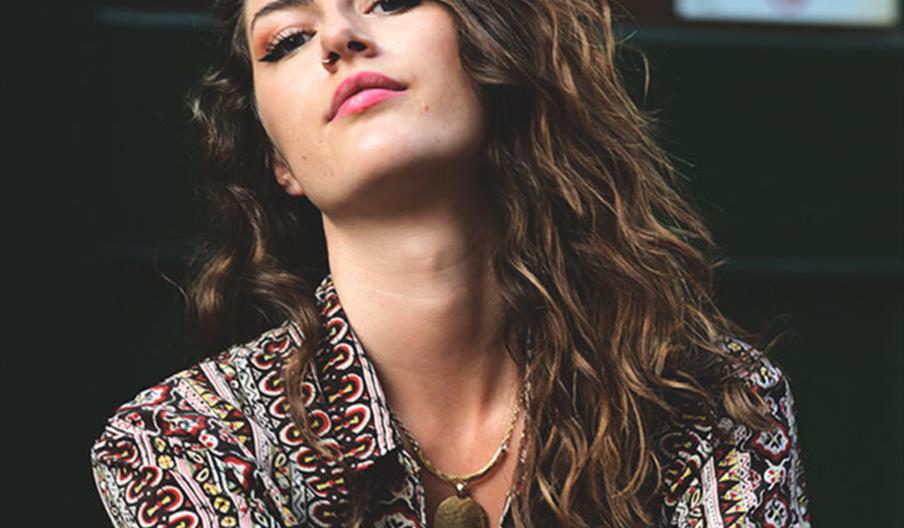 Concert poster featuring a young woman with wavy brown hair and a patterned shirt, looking confidently at the camera. Text announces "Lucca Mae" perfo