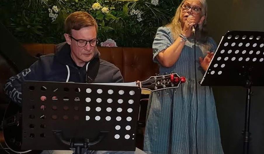 A man plays guitar while sitting behind a music stand, and a woman in a blue dress stands beside him singing into a microphone; both have music stands