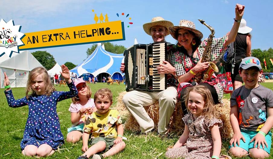 A group of children and two adults, one playing an accordion and the other a saxophone, sit on hay bales outdoors at a festival with tents in the back