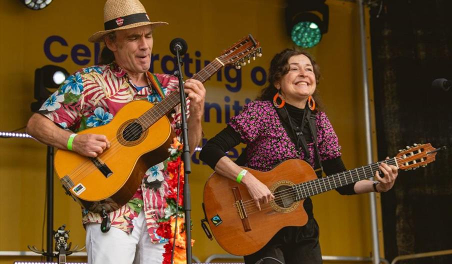 A man in a straw hat and a woman with large orange earrings are playing acoustic guitars and smiling while performing on an outdoor stage with a yello