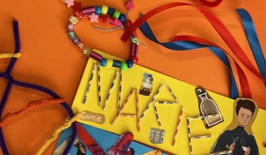 A colourful craft scene with beaded bracelets, pipe cleaners, red and blue ribbons, and a yellow card decorated with "Marie" stitched in yarn and illu