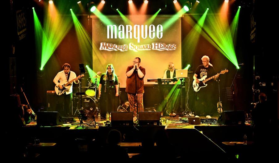 A band of six musicians performs on a stage under vibrant green and yellow lights, with instruments including electric guitars, a keyboard, drums, and