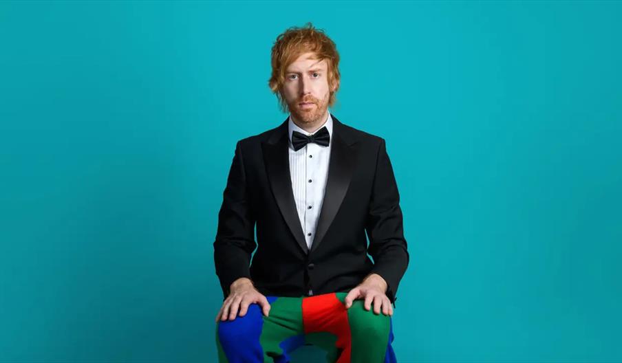 A man with light brown hair and beard, dressed in a formal black tuxedo and bow tie, sits against a teal background with his hands resting on his knee
