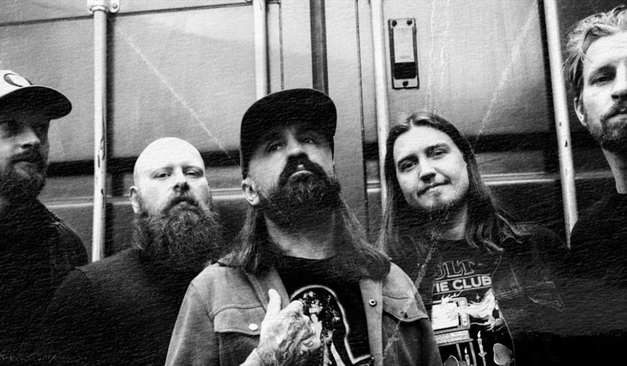 Black and white photo of five men with facial hair standing in front of a metal background, with serious and relaxed expressions, styled in casual or