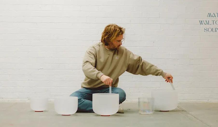 A person with shoulder-length hair and glasses sits cross-legged on the floor in front of a white brick wall, playing several crystal singing bowls ar