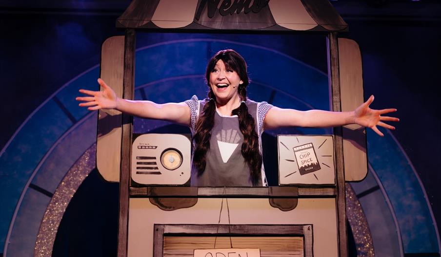 An actor holds their hands outward smiling dressed as Dorothy from The Wizard of Oz.