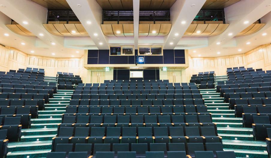 Interior of Middleton Hall. A wide view of an empty, modern lecture hall with tiered rows of dark blue seats, carpeted aisles, and well-lit wood-panel