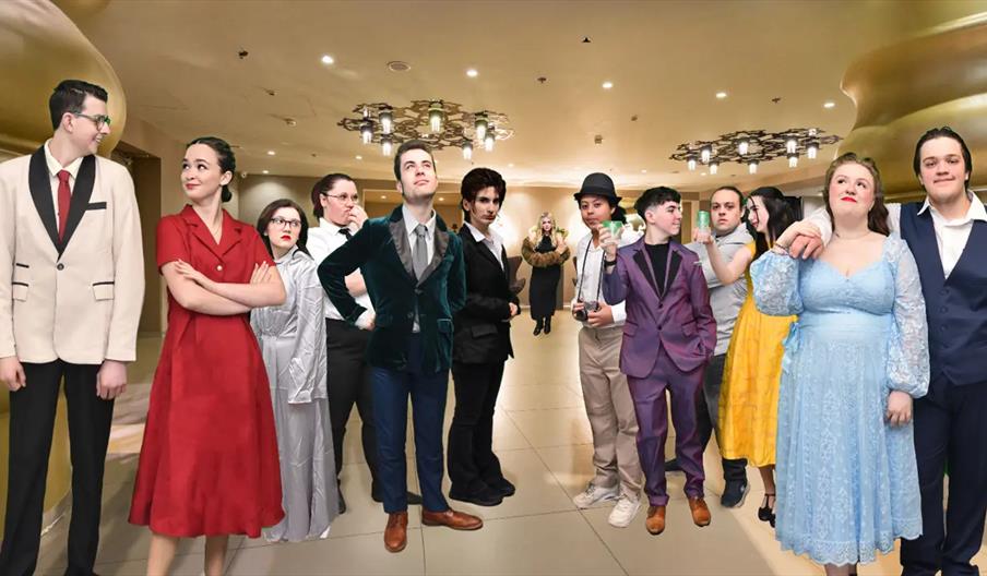 A group of thirteen people dressed in colorful formal and vintage-style attire stands in a well-lit hallway with modern chandeliers, posing together a