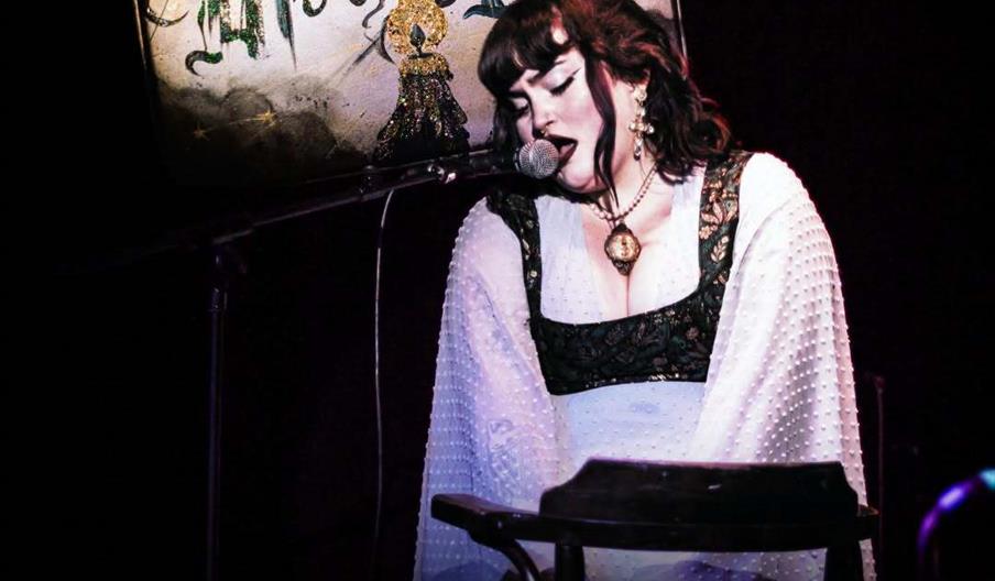 A woman in a white dress sings into a microphone on stage with a sign reading "Mira's Mar" behind her. Event details below her read: "Thursday 6th Aug