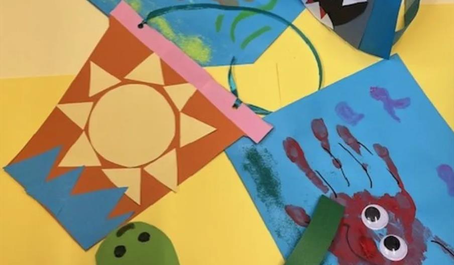 A collection of colorful children's craft projects, including a paper with a sun made of cut-out shapes, a blue sheet with a red handprint crab with g