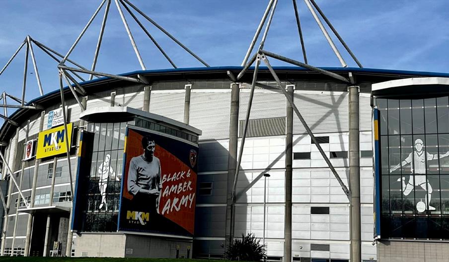 Exterior view of a modern stadium with a large sign reading "MKM Stadium," display banners showing a footballer and the text "Black & Amber Army," and