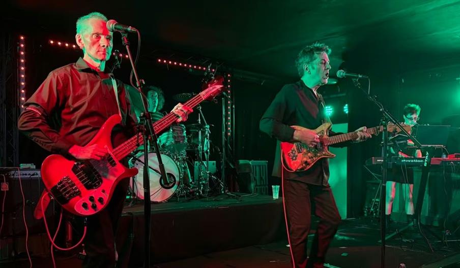 A four-member band performs on stage under green and red lighting, with musicians playing electric bass, electric guitar, drums, and keyboard.