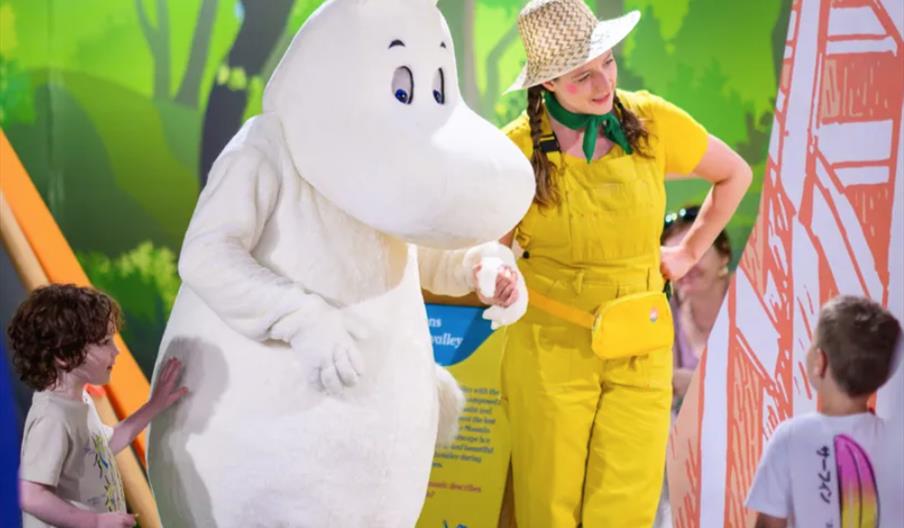 A person in a large Moomin character costume stands next to a woman dressed in a yellow outfit and straw hat, engaging with two young children in a co